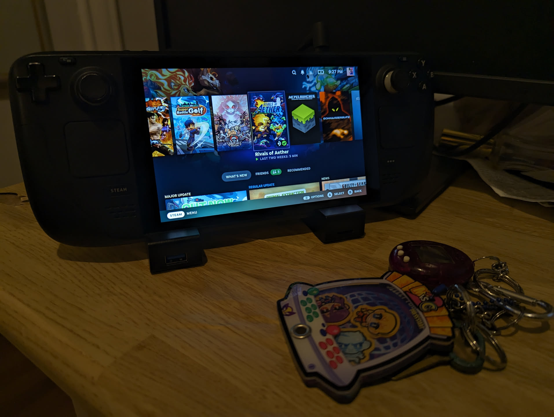 A picture of a Steam Deck, turned on, next to a keychain that has a Tamogatchi and a mini arcade cabinet attached to it.